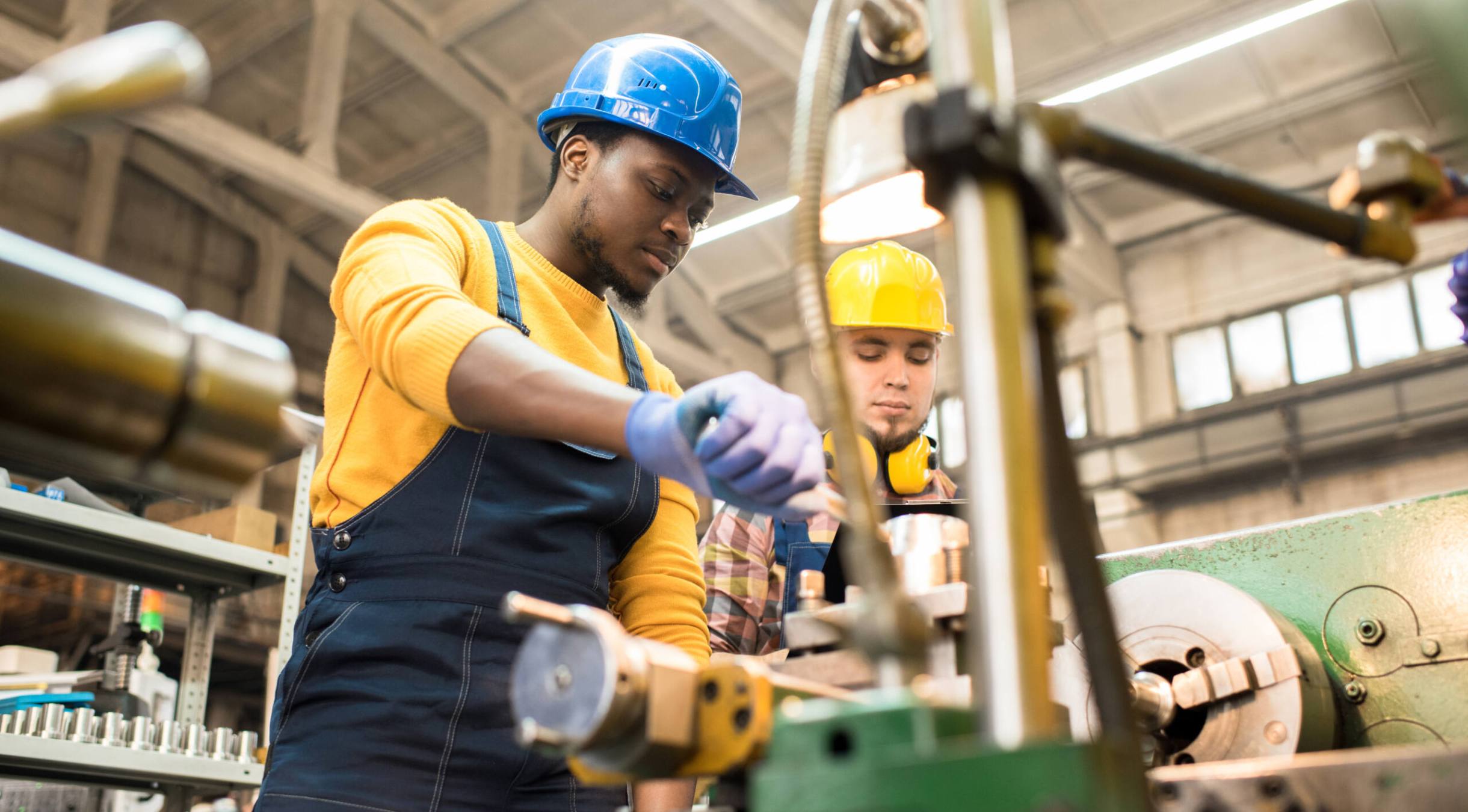Multi-ethnic team of workers wearing overalls and protective helmets using lathe in order to machine workpiece, interior of spacious production department on background